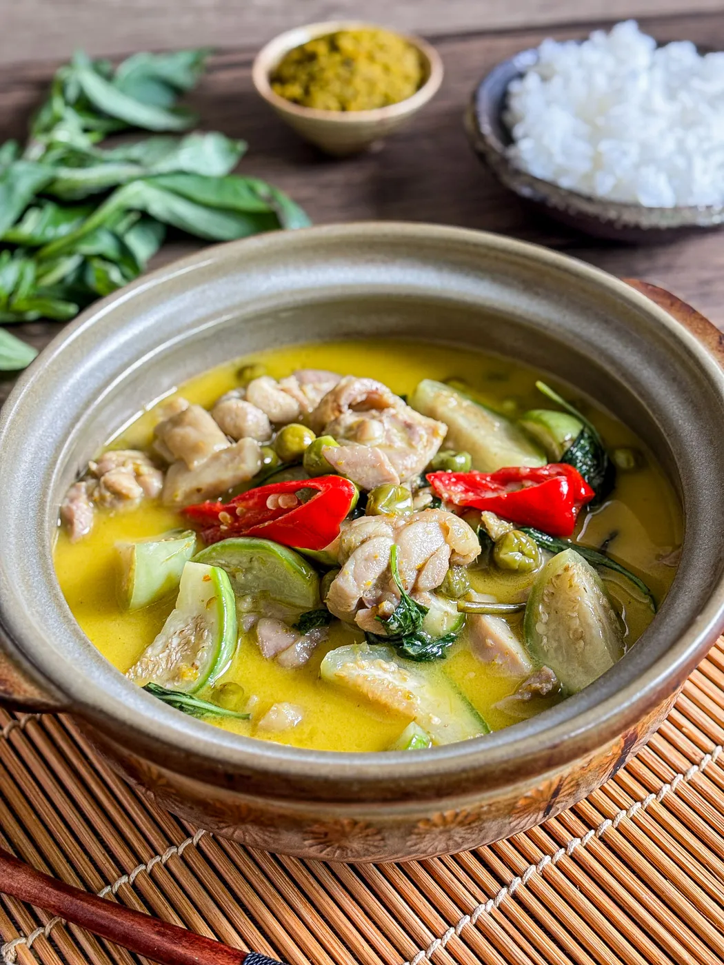 Authentic thai green curry with chicken and vegetables in a rich coconut broth, styled with Thai basil, red chili, and green curry paste behind the bowl.