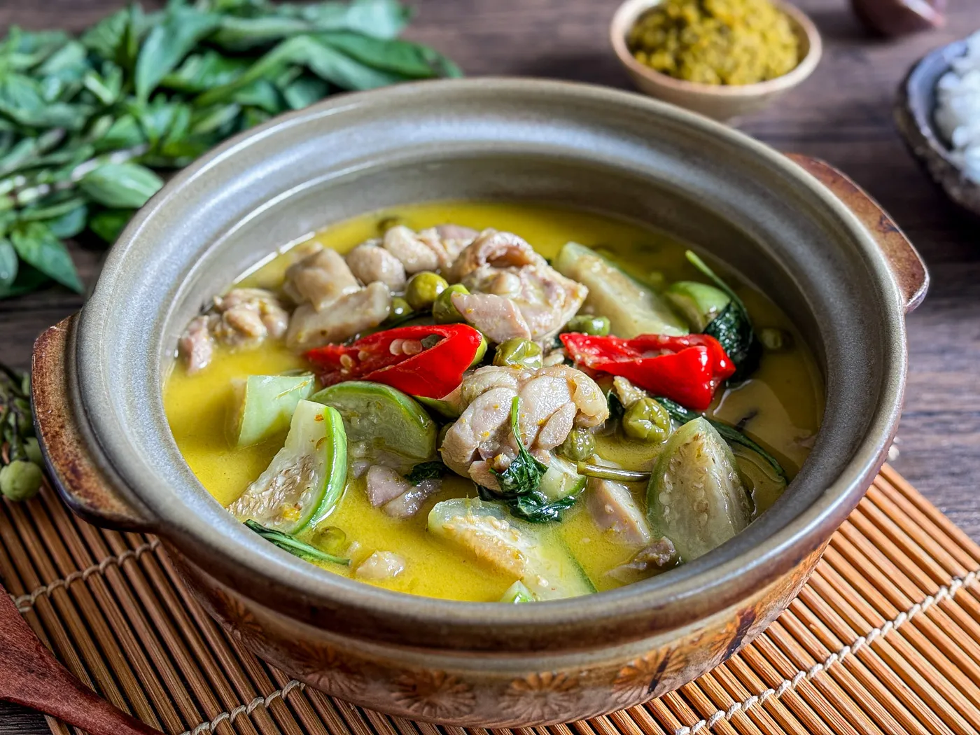 Authentic Thai green curry chicken with vegetables, coconut milk, and chilies served in a clay pot, with fresh basil and curry paste in the background.