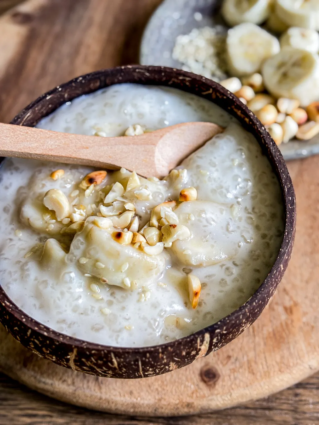 Close-up of Vietnamese banana tapioca pudding (chè chuối) served in a coconut bowl, topped with toasted peanuts and sesame seeds.