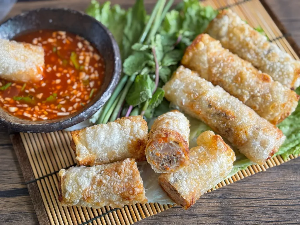 Cha gio arranged on a bamboo mat with fresh herbs and a garlic chili dipping sauce; one spring roll is dipped into the sauce in a black bowl.