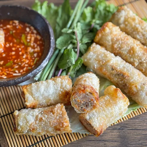 Cha gio arranged on a bamboo mat with fresh herbs and a garlic chili dipping sauce; one spring roll is dipped into the sauce in a black bowl.