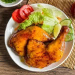 Filipino chicken inasal served with fresh lettuce, sliced tomatoes, cucumbers, and lime on a white plate, with annatto oil in the background.