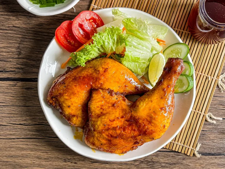 Filipino chicken inasal served with fresh lettuce, sliced tomatoes, cucumbers, and lime on a white plate, with annatto oil in the background.
