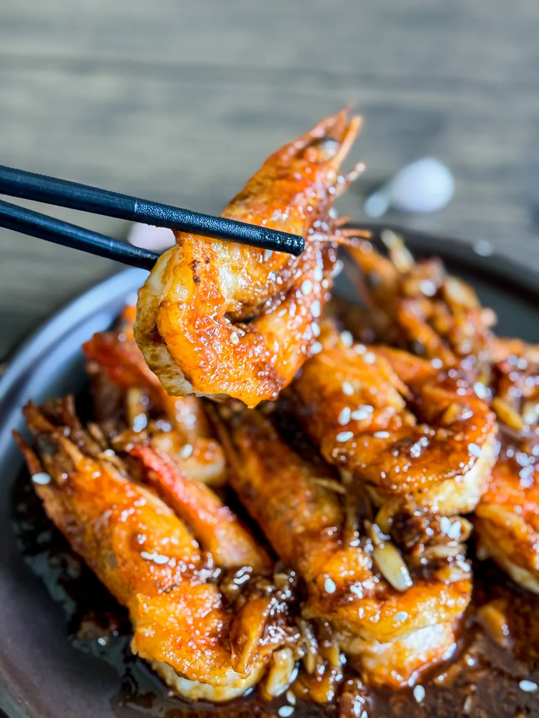 Close-up of chopsticks holding a crispy garlic shrimp with visible garlic pieces and sesame seeds on a dark serving plate.