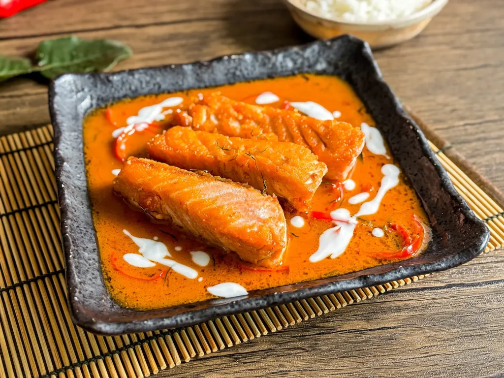 Pan-seared salmon fillets in choo chee curry sauce with coconut milk drizzle, served in a square black bowl with jasmine rice.