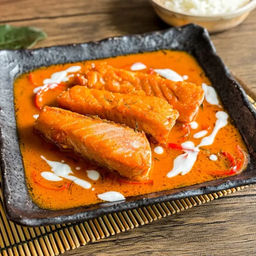 Pan-seared salmon fillets in choo chee curry sauce with coconut milk drizzle, served in a square black bowl with jasmine rice.