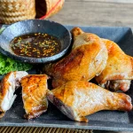 Gai yang, Thai grilled chicken served on a black plate with dipping sauce and lettuce on the side.