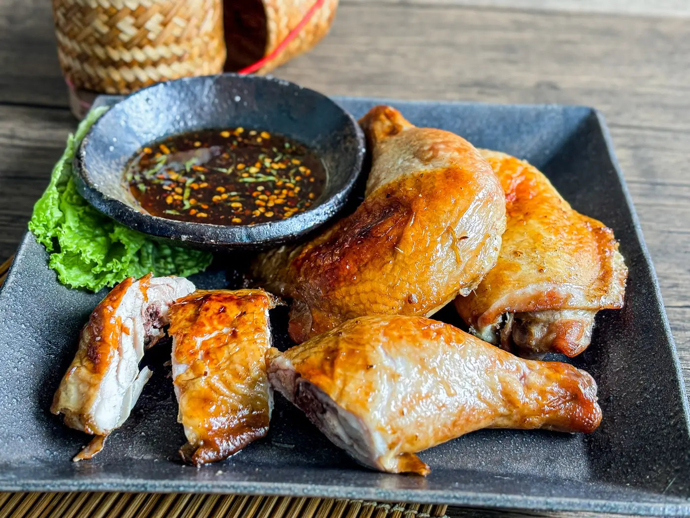 Gai yang, Thai grilled chicken served on a black plate with dipping sauce and lettuce on the side.