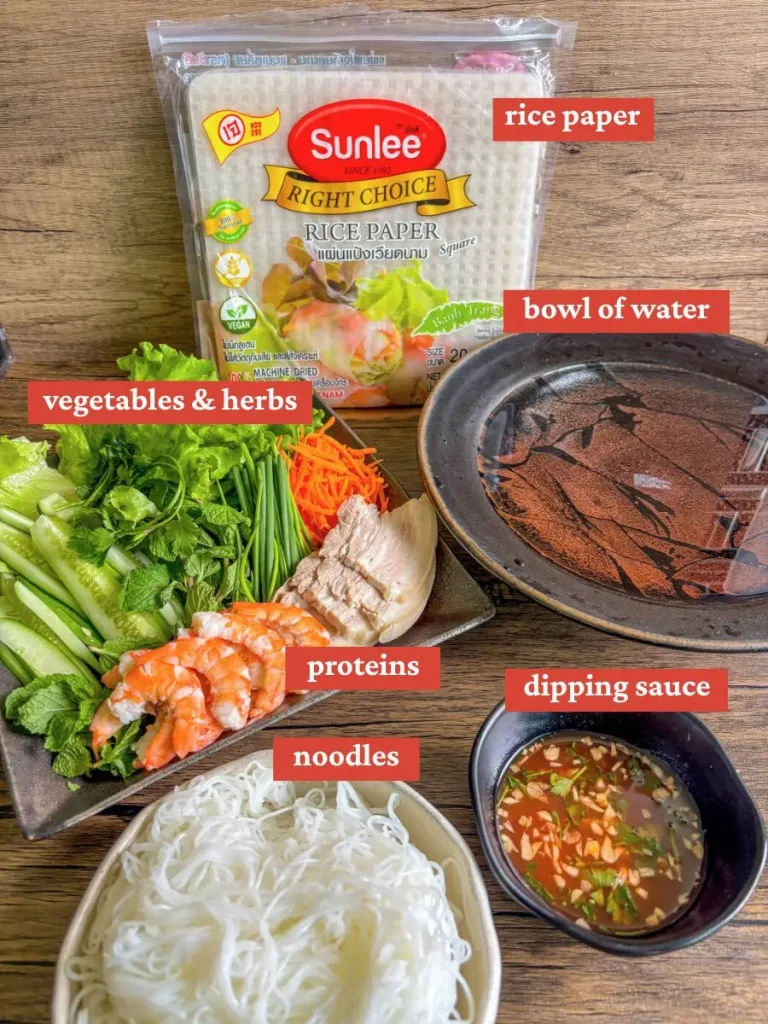 Flat lay of goi cuon ingredients including rice paper, shrimp, pork, vegetables, herbs, rice noodles, dipping sauce, and a bowl of water for softening wrappers.