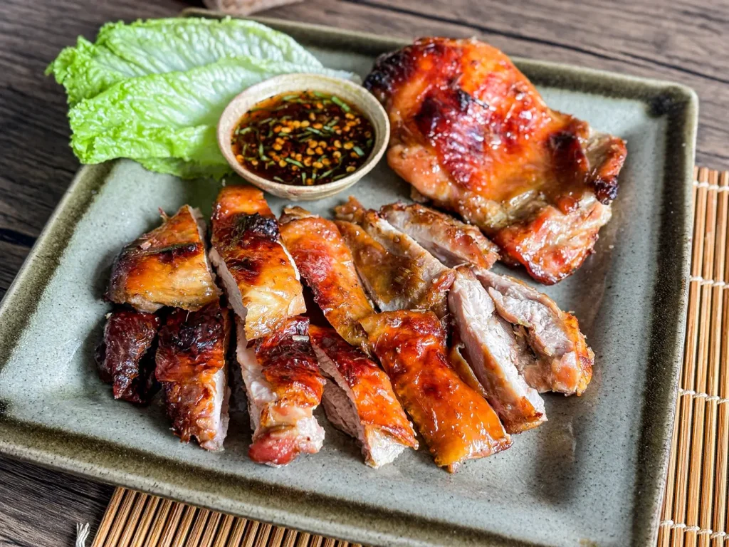 Thai grilled lemongrass chicken sliced and served with dipping sauce and lettuce on a ceramic plate.