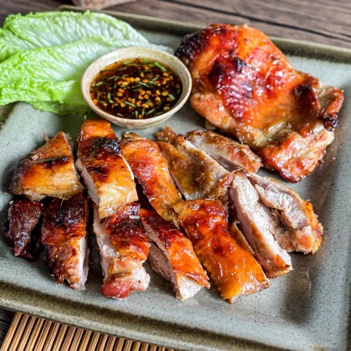 Thai grilled lemongrass chicken sliced and served with dipping sauce and lettuce on a ceramic plate.