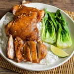 Hoisin chicken thighs served with rice, baby bok choy, and a lime wedge on a wooden mat.