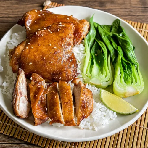 Hoisin chicken thighs served with rice, baby bok choy, and a lime wedge on a wooden mat.