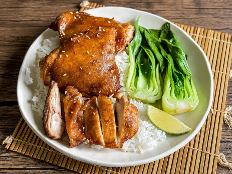 Hoisin chicken thighs served with rice, baby bok choy, and a lime wedge on a wooden mat.