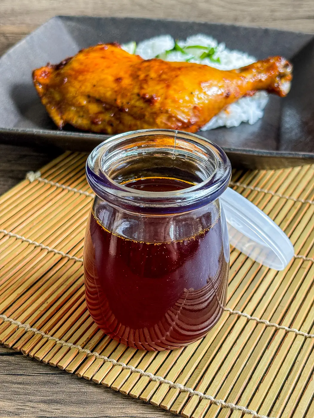 Filipino inasal chicken oil in a glass jar served with grilled chicken leg and rice in the background.