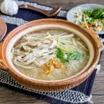 Khao tom gai in a rustic clay pot, with shredded chicken, ginger, scallions, and fresh herbs on top, served with garnishes on the side.