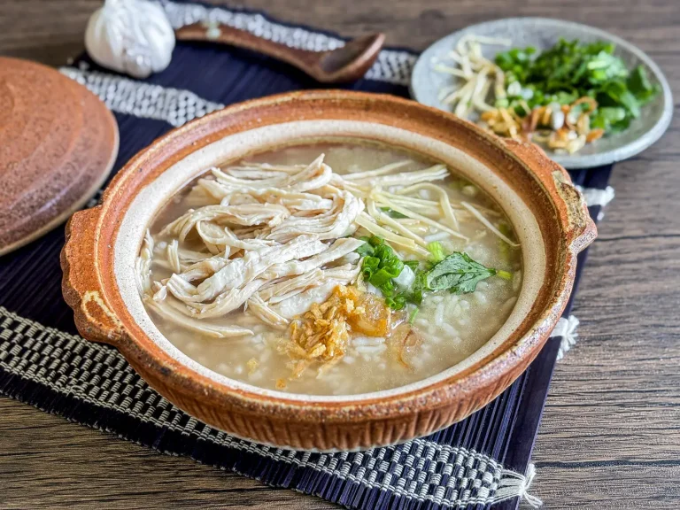 Khao tom gai in a rustic clay pot, with shredded chicken, ginger, scallions, and fresh herbs on top, served with garnishes on the side.
