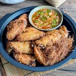 Thai lemongrass fried chicken wings served in a black plate, topped with crispy lemongrass threads and a side of sweet chili sauce.