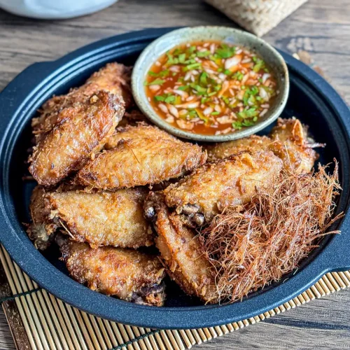 Thai lemongrass fried chicken wings served in a black plate, topped with crispy lemongrass threads and a side of sweet chili sauce.