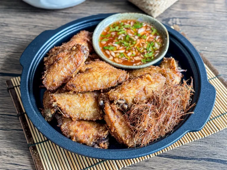 Thai lemongrass fried chicken wings served in a black plate, topped with crispy lemongrass threads and a side of sweet chili sauce.