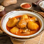 Thai massaman curry with chicken drumstick, golden potatoes, peanuts, and onions in rich red coconut curry sauce, with rice and spices in the background.