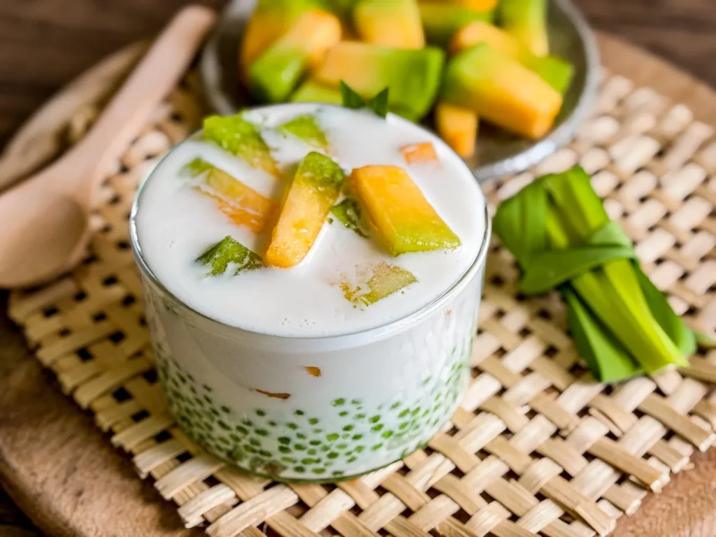 Glass filled with coconut melon sago dessert topped with green and orange melon slices, served with pandan leaves and a wooden spoon.