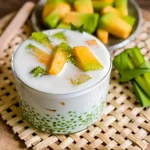 Glass filled with coconut melon sago dessert topped with green and orange melon slices, served with pandan leaves and a wooden spoon.