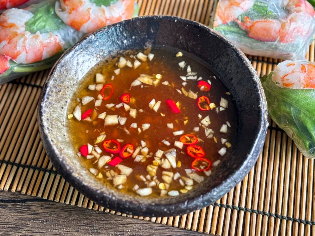Nuoc cham, a Vietnamese fish dipping sauce, in a dark ceramic bowl, topped with minced garlic and sliced red chili, served with fresh spring rolls.