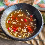 Nuoc cham, a Vietnamese fish dipping sauce, in a dark ceramic bowl, topped with minced garlic and sliced red chili, served with fresh spring rolls.