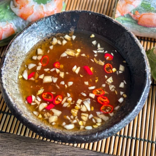 Nuoc cham, a Vietnamese fish dipping sauce, in a dark ceramic bowl, topped with minced garlic and sliced red chili, served with fresh spring rolls.