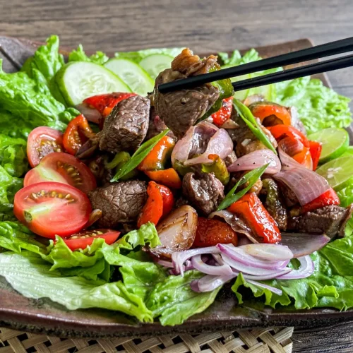Chopsticks holding a piece of shaking beef over a lettuce-lined plate filled with sautéed beef, red onions, tomatoes, and fresh lime wedges.
