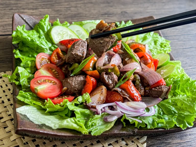 Chopsticks holding a piece of shaking beef over a lettuce-lined plate filled with sautéed beef, red onions, tomatoes, and fresh lime wedges.