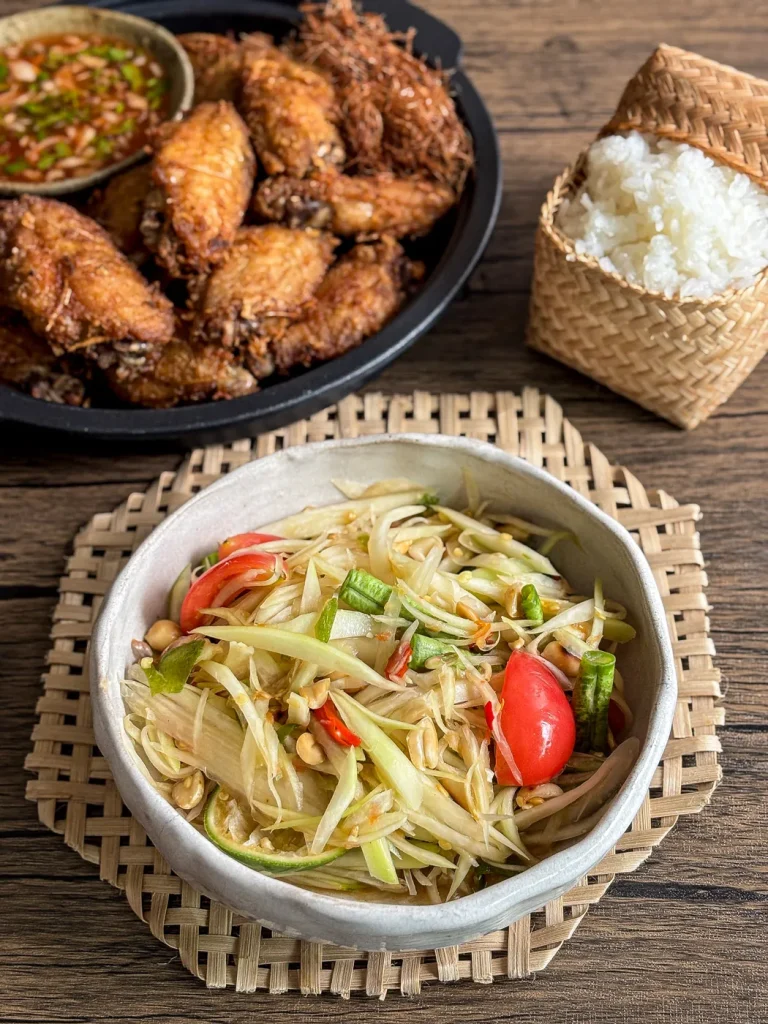 Som tam (Thai papaya salad) served in a white bowl with tomatoes, lime, and peanuts, paired with sticky rice and crispy fried chicken in the background.