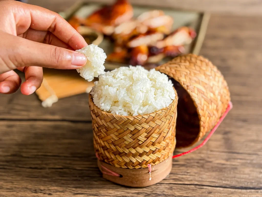 A hand picking up Lao sticky rice from a traditional bamboo basket, with grilled meat and sauce blurred in the background.