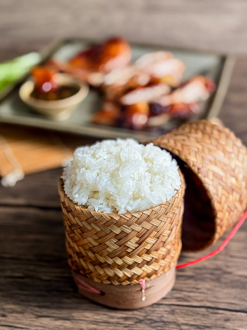 Freshly steamed Thai sticky rice served in a traditional bamboo basket, with grilled chicken and dipping sauce in the background.