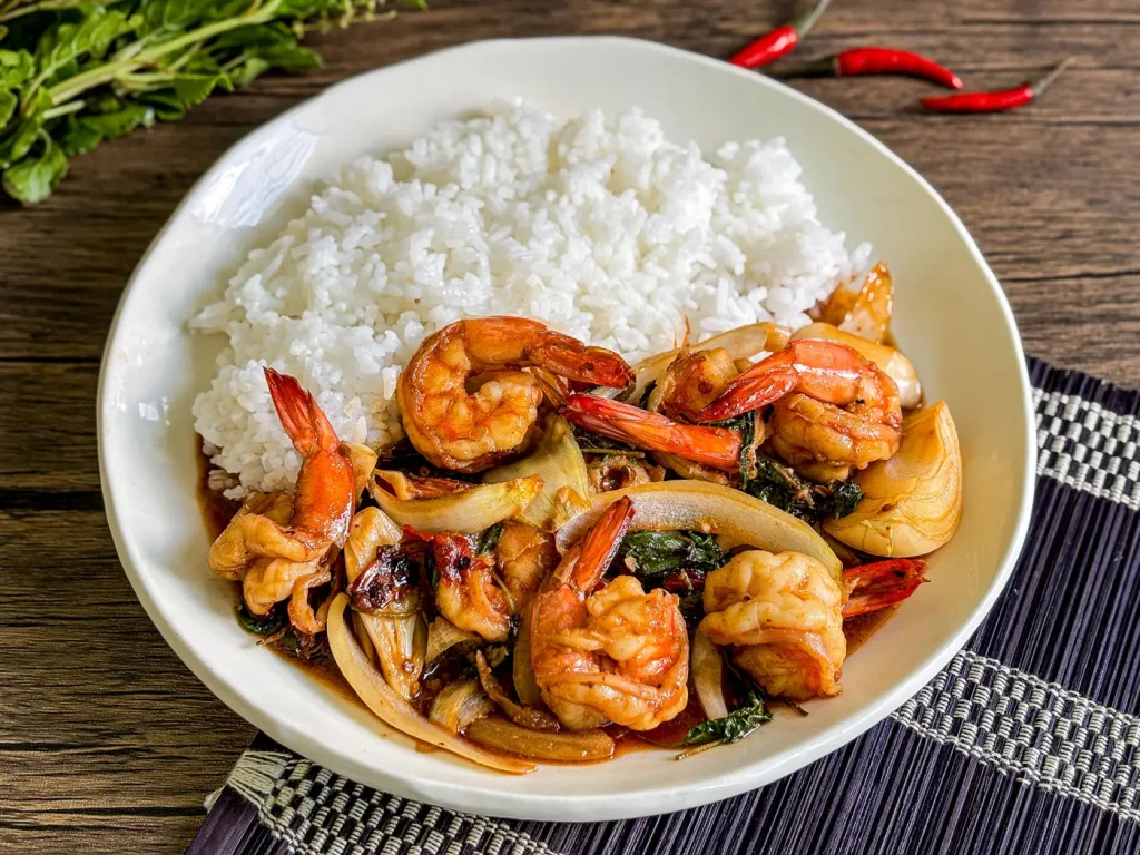 Thai basil shrimp stir-fry served with jasmine rice on a white plate, styled with fresh red chilies and holy basil on a wooden table.