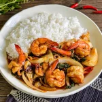 Thai basil shrimp stir-fry served with jasmine rice on a white plate, styled with fresh red chilies and holy basil on a wooden table.