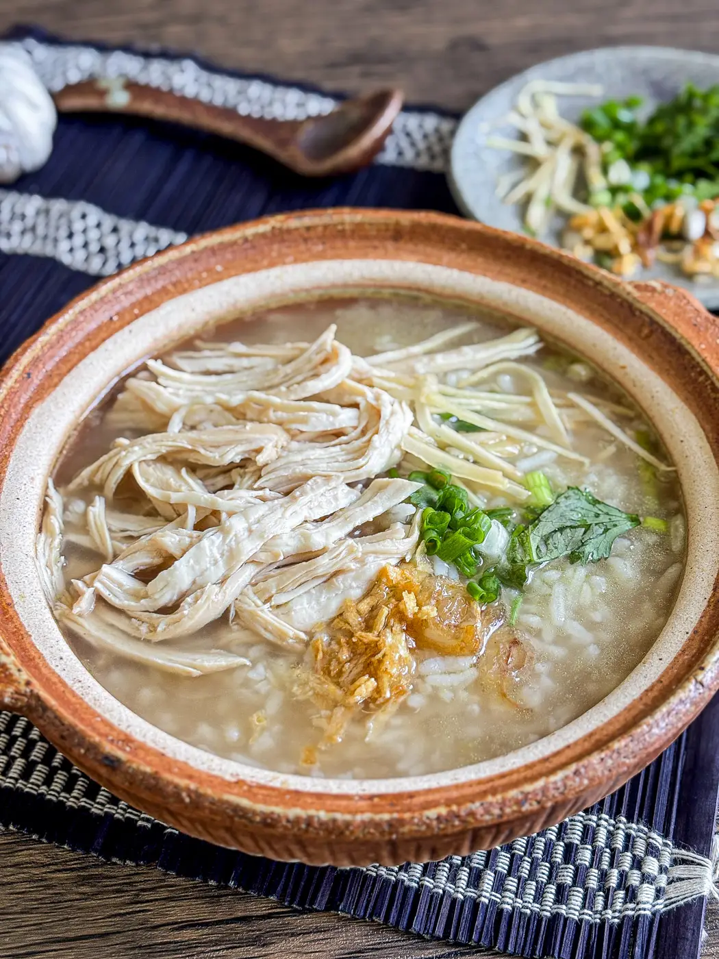 Clay bowl of Thai chicken and rice soup topped with shredded chicken, fried garlic, scallions, and herbs, served on a woven placemat.