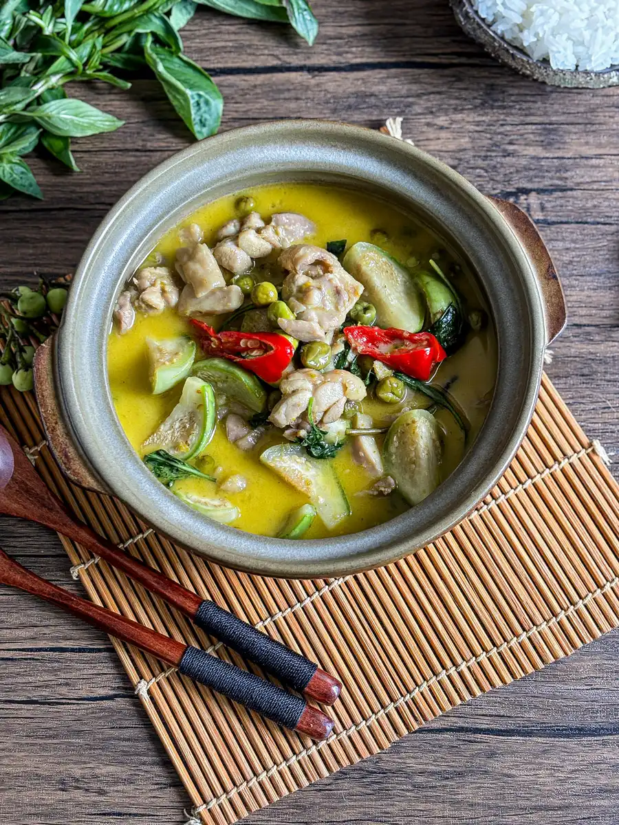 Thai green chicken curry in a clay pot with Thai eggplants, pea eggplants, red chili, and basil, served with jasmine rice on a bamboo mat.