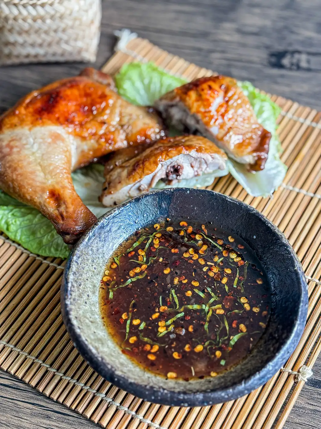 Thai grilled chicken dipping sauce served in a rustic bowl, topped with chili flakes and herbs, with grilled chicken leg in the background.