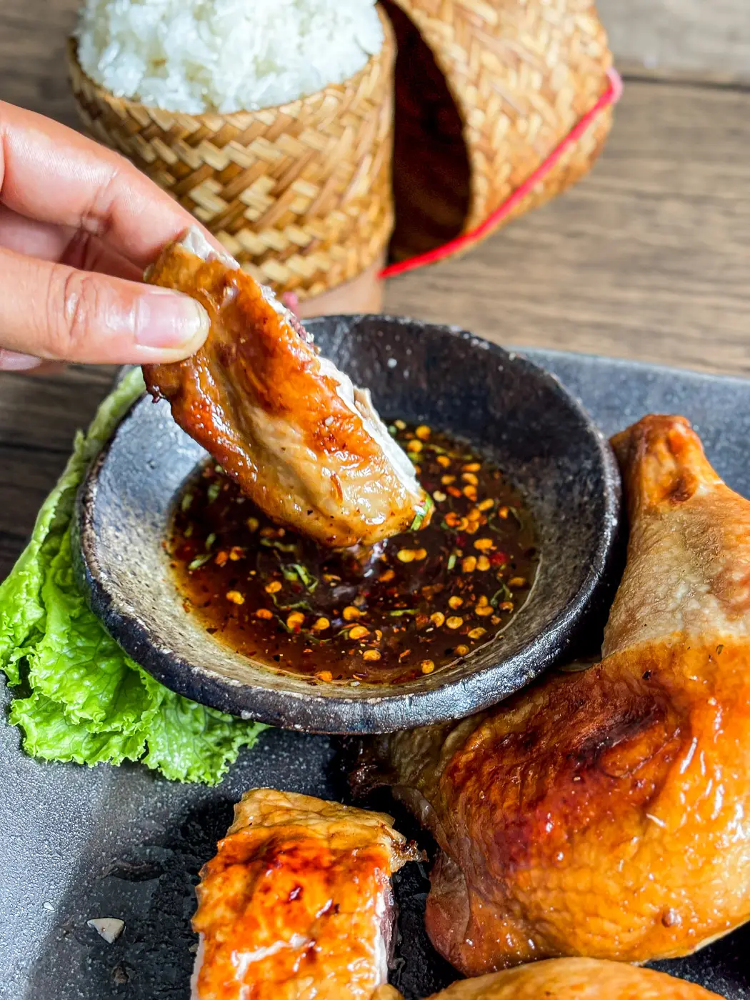 A piece of Thai grilled chicken being dipped into spicy dipping sauce, with sticky rice in a bamboo basket in the background.