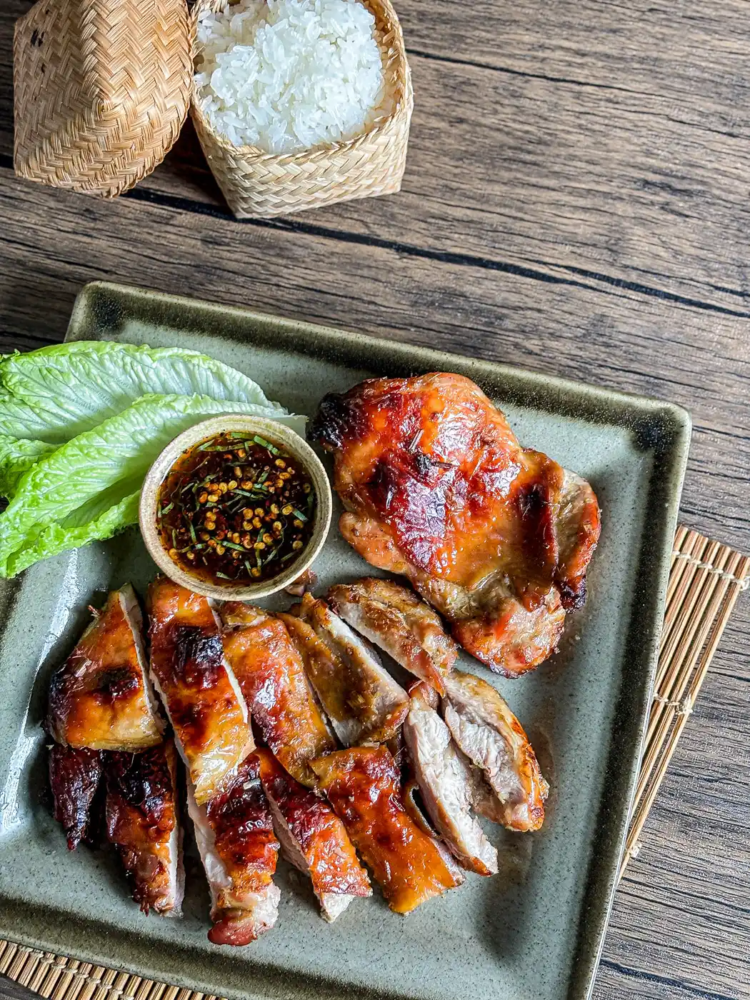 Overhead view of Thai grilled lemongrass chicken with sticky rice in a bamboo basket, served with dipping sauce and fresh lettuce.