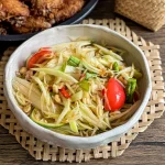 Thai papaya salad (som tam) in a ceramic bowl, topped with tomatoes, chilies, long beans, and peanuts, with sticky rice and fried chicken.