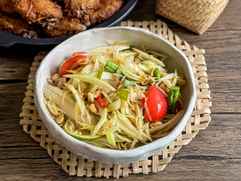 Thai papaya salad (som tam) in a ceramic bowl, topped with tomatoes, chilies, long beans, and peanuts, with sticky rice and fried chicken.