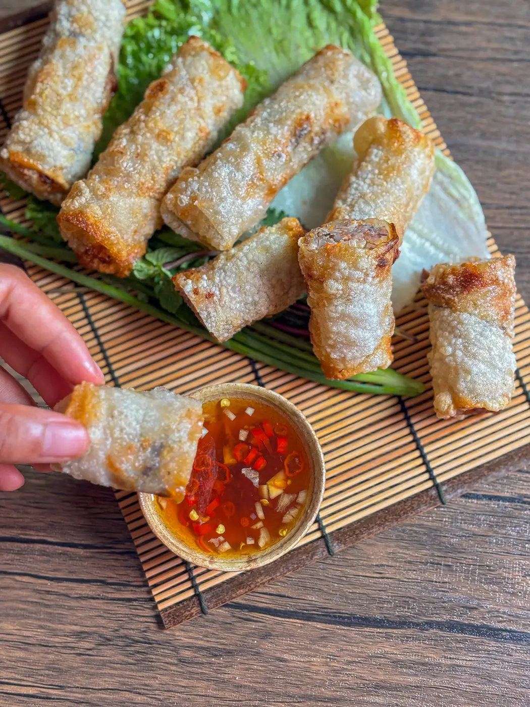 Vietnamese fried spring rolls served on lettuce with a dipping bowl of nuoc cham; one roll is being dipped by hand into the sauce.