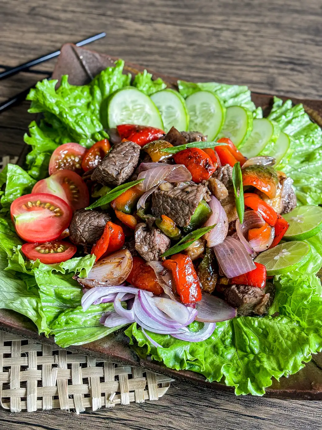 Vietnamese shaking beef with seared beef cubes, bell peppers, and onions, served on a bed of lettuce with cucumber and tomato slices.
