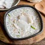 Thai coconut sago pudding served in a dark ceramic bowl, topped with slices of young coconut, sesame seeds, and pandan leaf on a wooden background.
