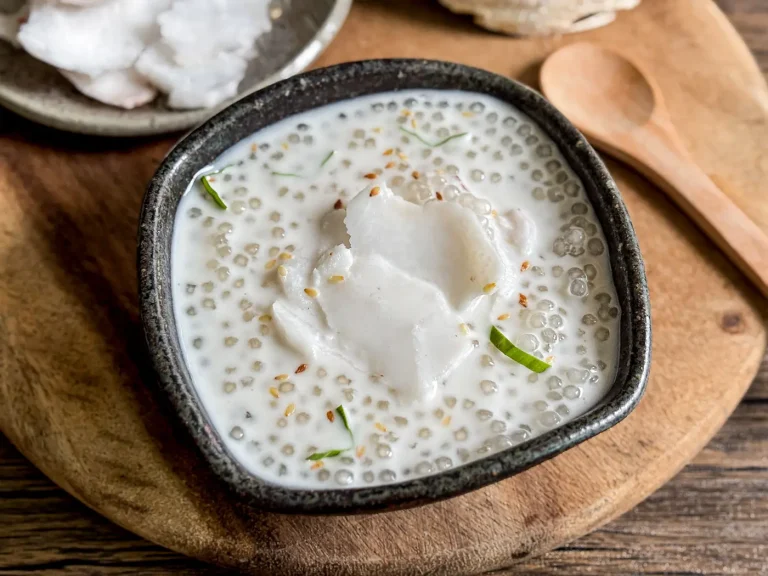 Thai coconut sago pudding served in a dark ceramic bowl, topped with slices of young coconut, sesame seeds, and pandan leaf on a wooden background.