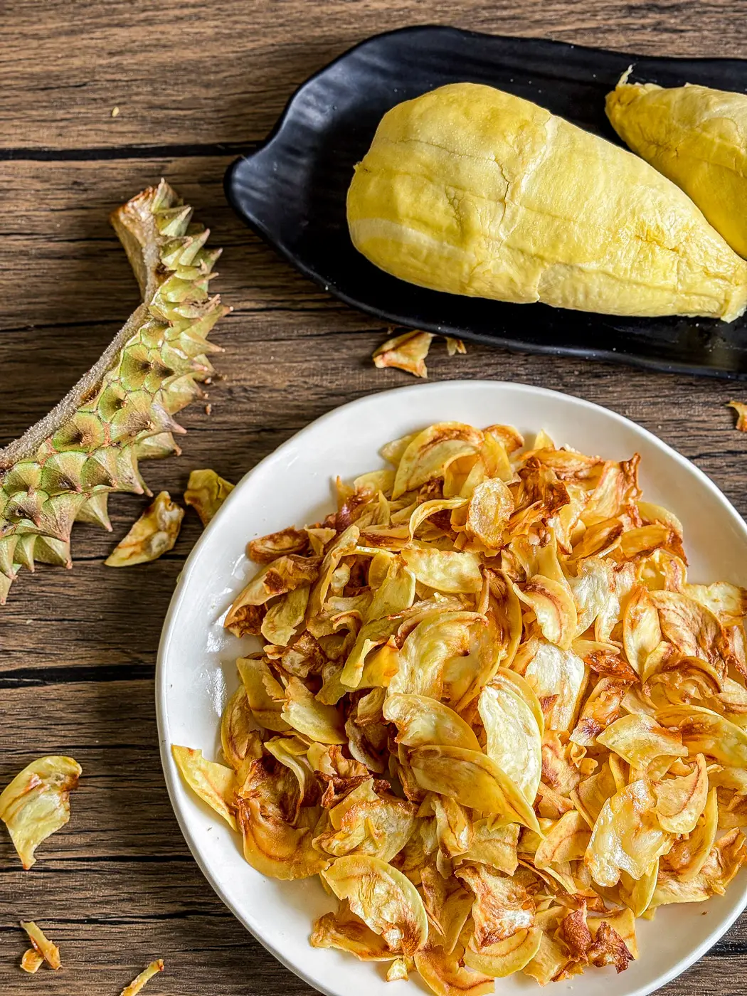 Crispy durian chips on white plate with durian fruit and spiky husk on rustic wooden table.