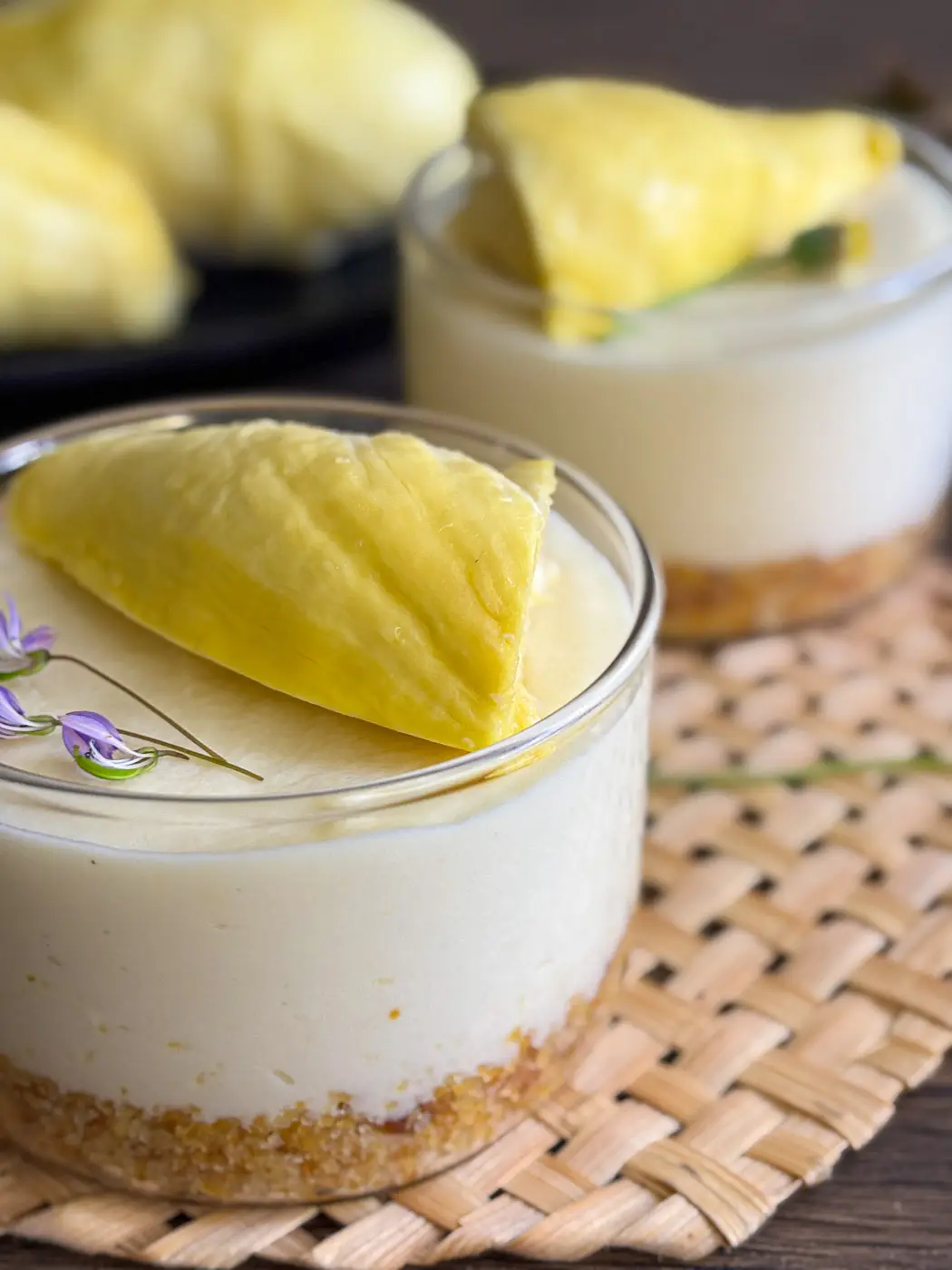 Close-up of durian cheesecake in a glass topped with a ripe durian chunk and edible flowers, showing smooth texture and crumb crust.